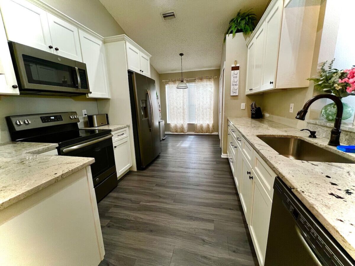Una cocina con gabinetes blancos, microondas, estufa, refrigerador y lavavajillas.