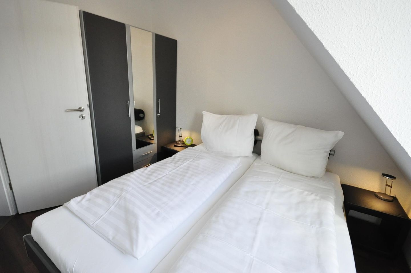 A single bed with white bedding in a room with a sloped ceiling, a wardrobe, a bedside table with a lamp, and a small clock.