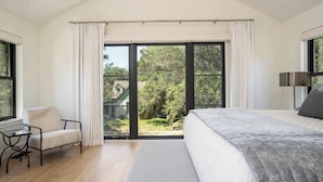 Quarto com uma cama grande, uma cadeira, uma mesa e vista para um jardim através de uma porta deslizante.