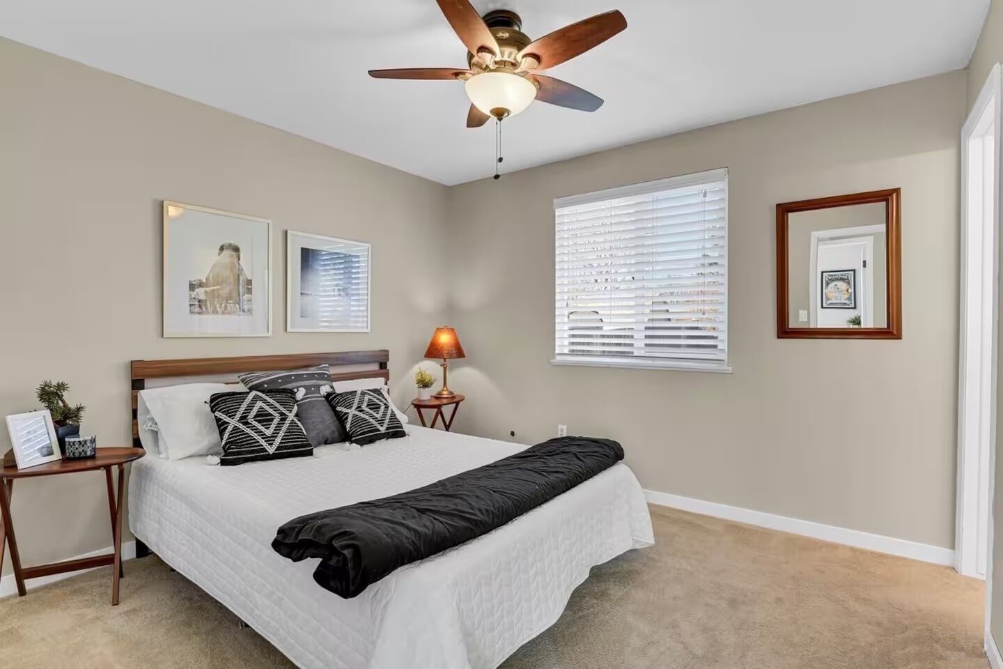 A bedroom with a bed, a ceiling fan, a nightstand with a lamp, and framed pictures on the wall.