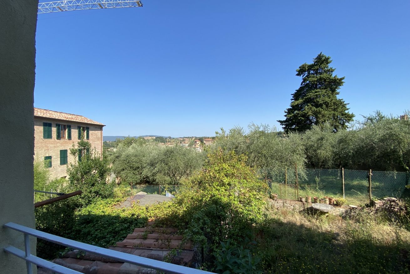 A courtyard with a view of a building with green shutters, a tall tree, and a fenced area with plants.