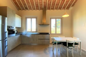 A modern kitchen with a central island, stainless steel appliances, and a dining area with white chairs and a table. - TRA LE MURA – Downtown Apartment in Siena (Siena)