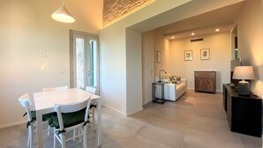 Ein modernes Wohnzimmer mit einem weißen Esstisch, einer Couch und einem Couchtisch. Ein Fenster mit Jalousien, eine Lampe und gerahmte Bilder sind an der Wand zu sehen.