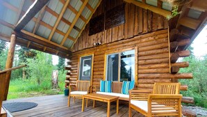 Une cabane en rondins dotée d’une terrasse en bois, d’un espace salon couvert avec des chaises et une table, et d’une grande fenêtre.