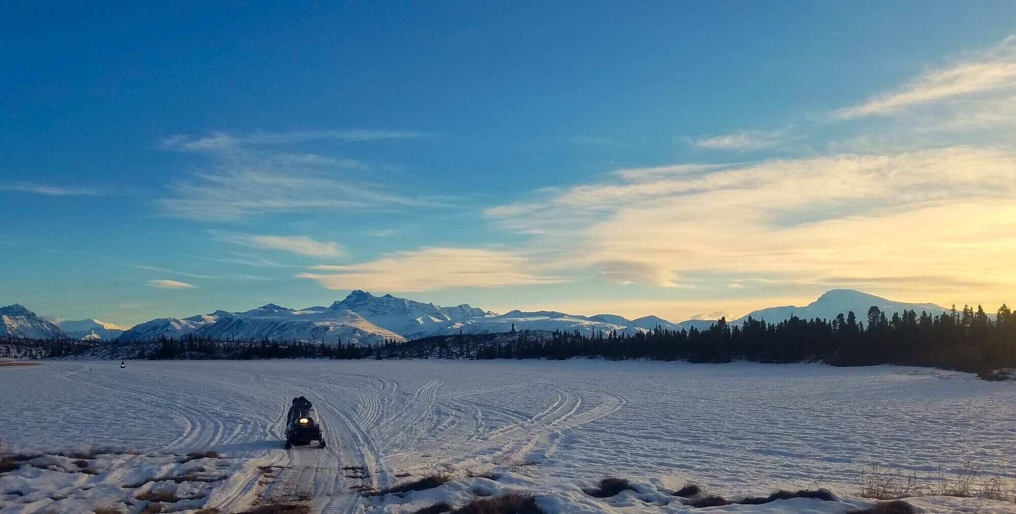 Paisaje nevado con un vehículo en un camino despejado, rodeado de montañas y un bosque.