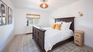 Ein Schlafzimmer mit einem dunklen Holzbett, weißer Bettwäsche, einem kleinen Nachttisch, einer Wandleuchte und einem Fenster mit einer Holzjalousie.