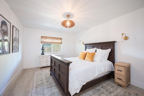 A bedroom with a dark wooden bed, white bedding, a small bedside table, a nightstand, a wall-mounted lamp, and a window with a wooden blind.