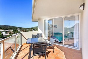 Un balcon avec une table et des chaises, un lit recouvert d’une couverture verte et une porte vitrée donnant sur une chambre à coucher.