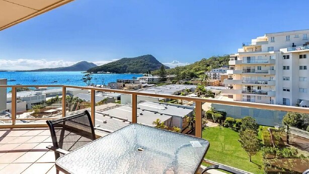 Ein Balkon mit Glastisch und Stühlen mit Blick auf eine Küstenlandschaft, einschließlich Gebäude und einem Berg.