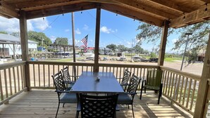A covered outdoor area with a wooden deck, a table with chairs, and a view of boats and a flag in the background.