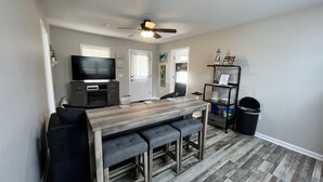 A compact living space with a wooden dining table, two grey cushioned benches, a television on a cabinet, a ceiling fan, and a small trash bin.