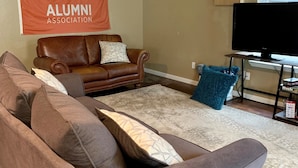 A living room with a brown leather sofa, a grey couch, and a flat-screen TV on a stand. A banner for the OSU Alumni Association is visible on the wall.
