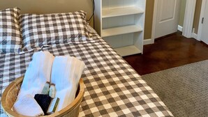 A bedroom with a checkered bedspread, a bedside lamp, a wicker basket with towels, and a white shelving unit.