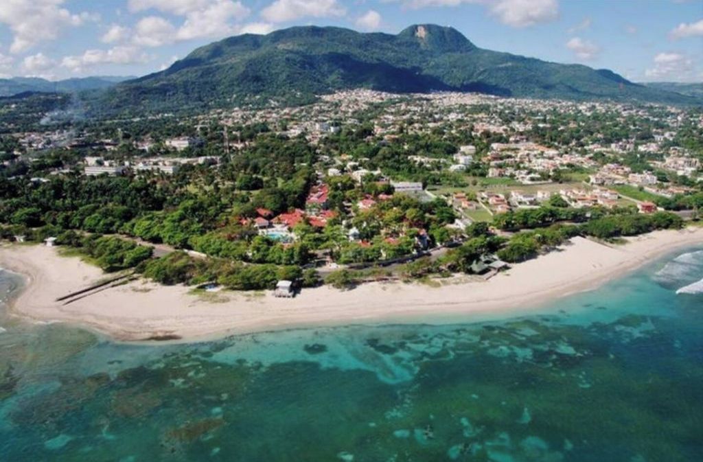 Luchtbeeld van een kustplaatsje met een strand, helder water en een bergachtige achtergrond.