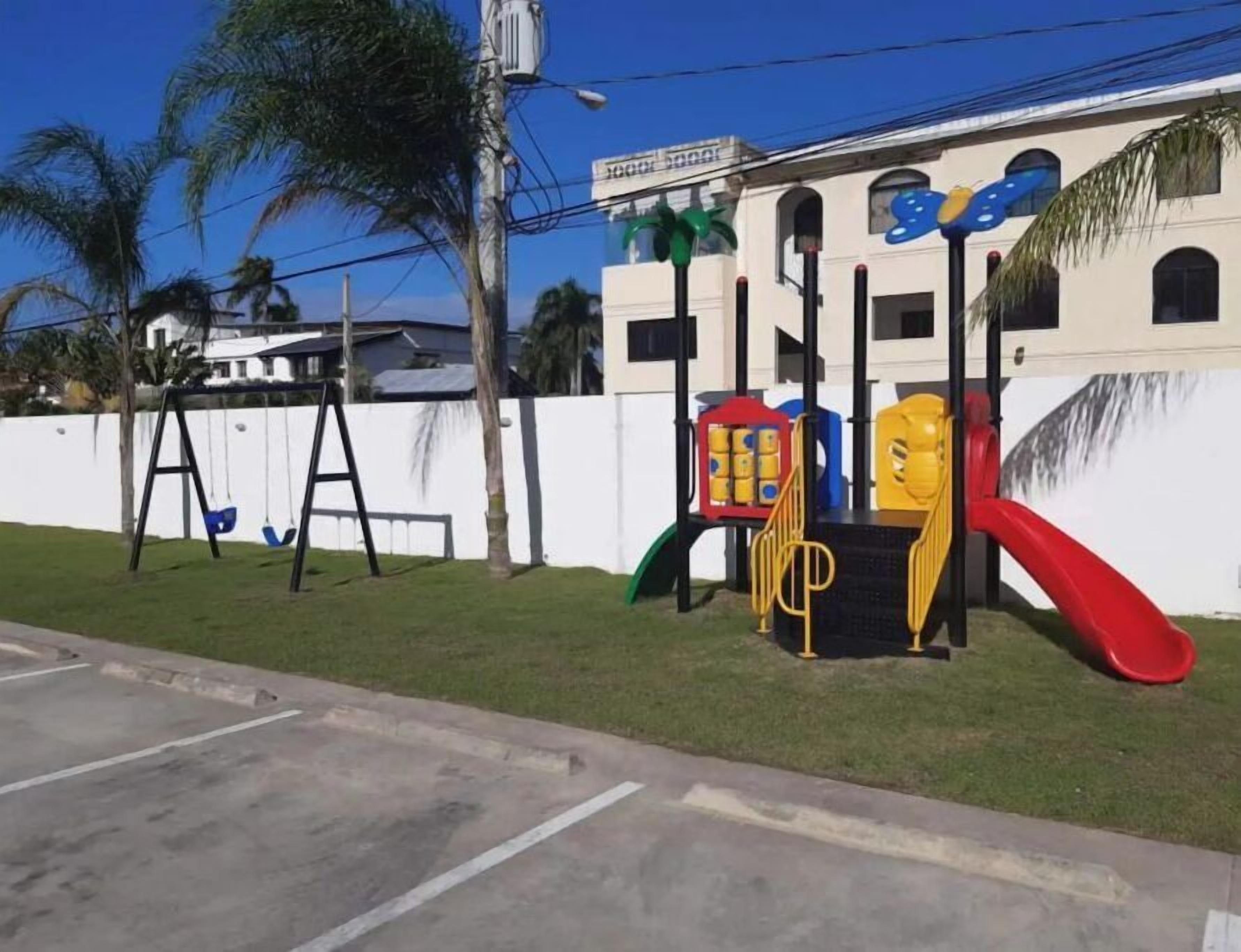 A playground with swings and a slide, a white fence, and a building with a sign in the background.