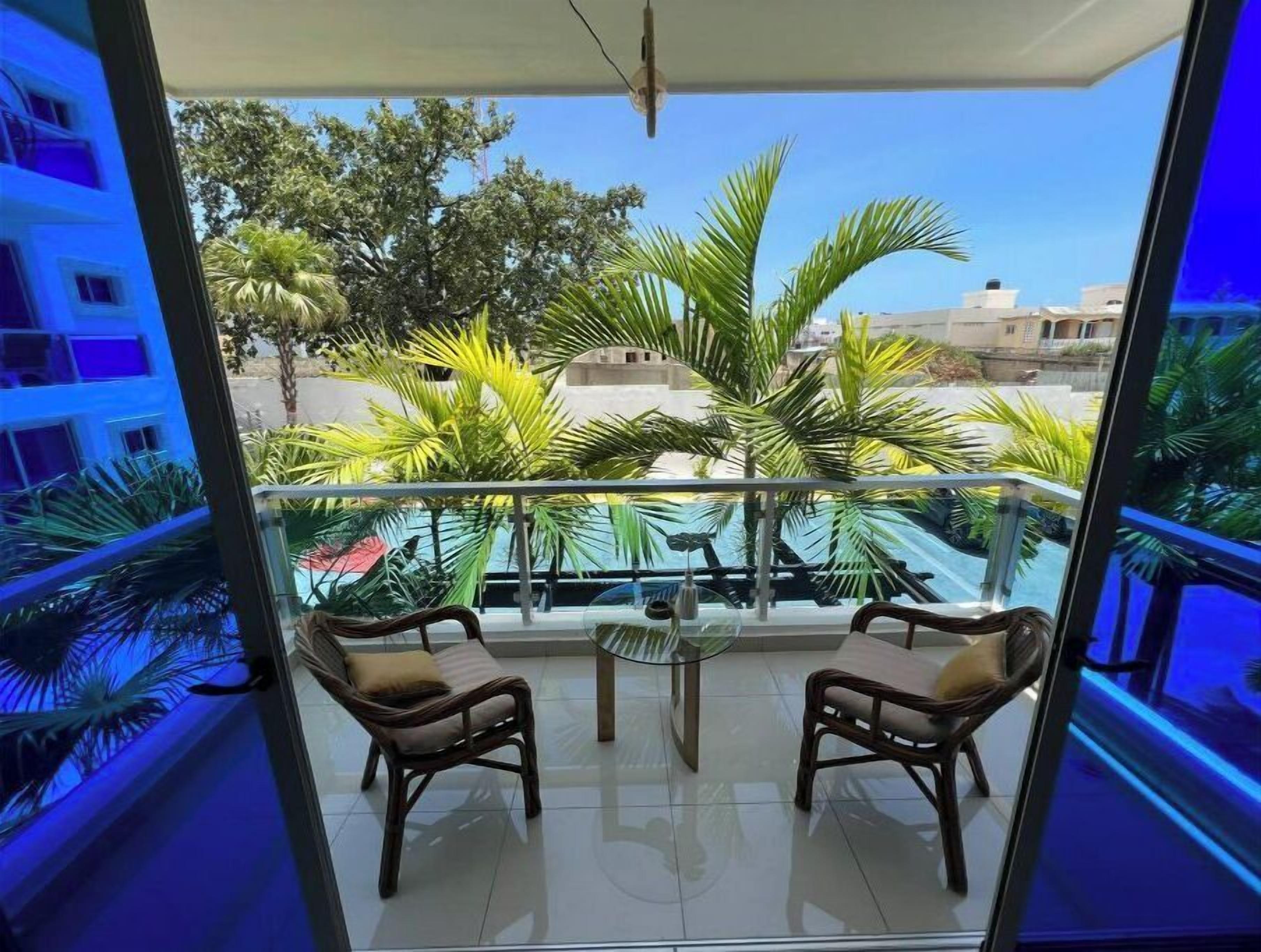 A balcony with two wicker chairs and a small table, overlooking a residential area with palm trees and houses.