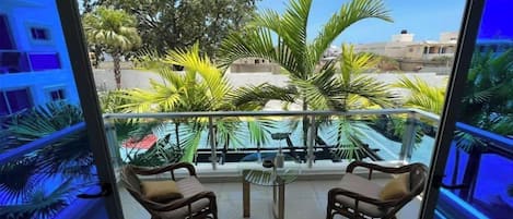 Un balcon avec deux fauteuils en osier et une petite table, donnant sur un quartier résidentiel bordé de palmiers et de maisons.