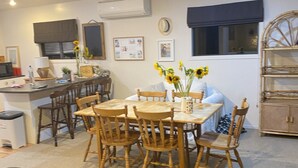 A dining area with a round table, wooden chairs, and a vase with sunflowers. There is a kitchen area with cabinets and a refrigerator in the background.