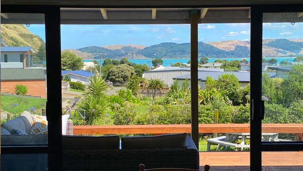 A balcony with a view of a lake and hills, wicker furniture, and a table.