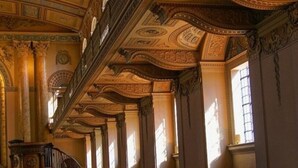 A grand hallway with ornate ceiling decorations and columns, leading to a large arched doorway.