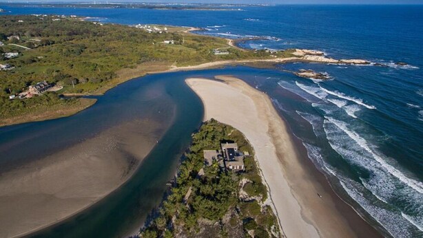 An aerial view of a coastal area with a beach, a small house, and surrounding greenery.