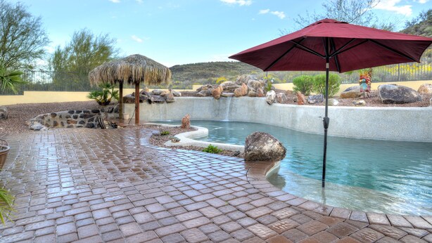 Uma piscina com um terraço de tijolos, um guarda-sol de palha e uma cascata.
