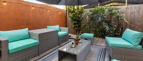An outdoor patio area with wicker furniture, a glass-top coffee table, and a large umbrella.