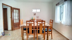 A dining area with a wooden table and chairs, a tiled floor, a door, and a window with curtains. - Casa do Romaninho (Porto da Cruz)