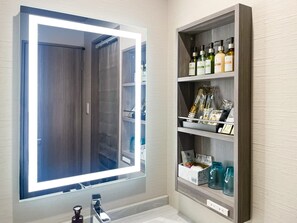 A bathroom with a large mirror, a vanity with a sink, and a shelf with toiletries.