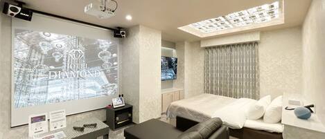A modern hotel room with a large screen displaying the hotel's name, a comfortable bed, a sofa, a small table with a speaker, and a ceiling light fixture.