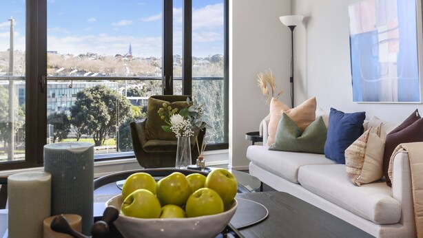 Una sala de estar moderna con un ventanal grande, un sofá con cojines de colores, un cuenco de manzanas y un jarr ón con flores.