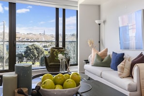 A modern living room with a large window, a sofa with cushions, a bowl of apples, and a vase with flowers.