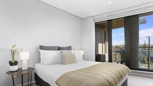 Un dormitorio moderno con una cama grande, una mesita de noche pequeña con una lámpara y vistas a la ciudad a través de amplias ventanas.