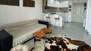 Una sala moderna con un sofá, dos taburetes altos y un tapiz de cuero de vaca. Se ve una cocina con taburetes blancos y un comedor al fondo.