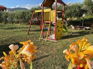 A playground with a wooden structure, including a slide and climbing area, surrounded by grass and flowering plants.