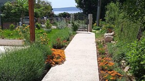 Un chemin en béton, bordé de différentes plantes et fleurs, qui mène à un portail.