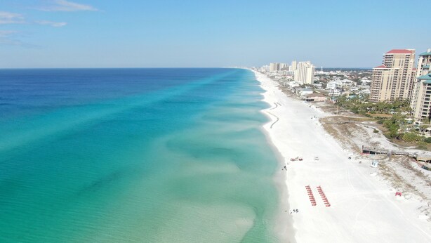 A coastal beach with a clear turquoise sea, white sand, and a row of buildings along the shore.