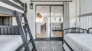 A room with two beds, a ladder, a patterned floor, a sliding glass door leading to a dining area, and a window with blinds.