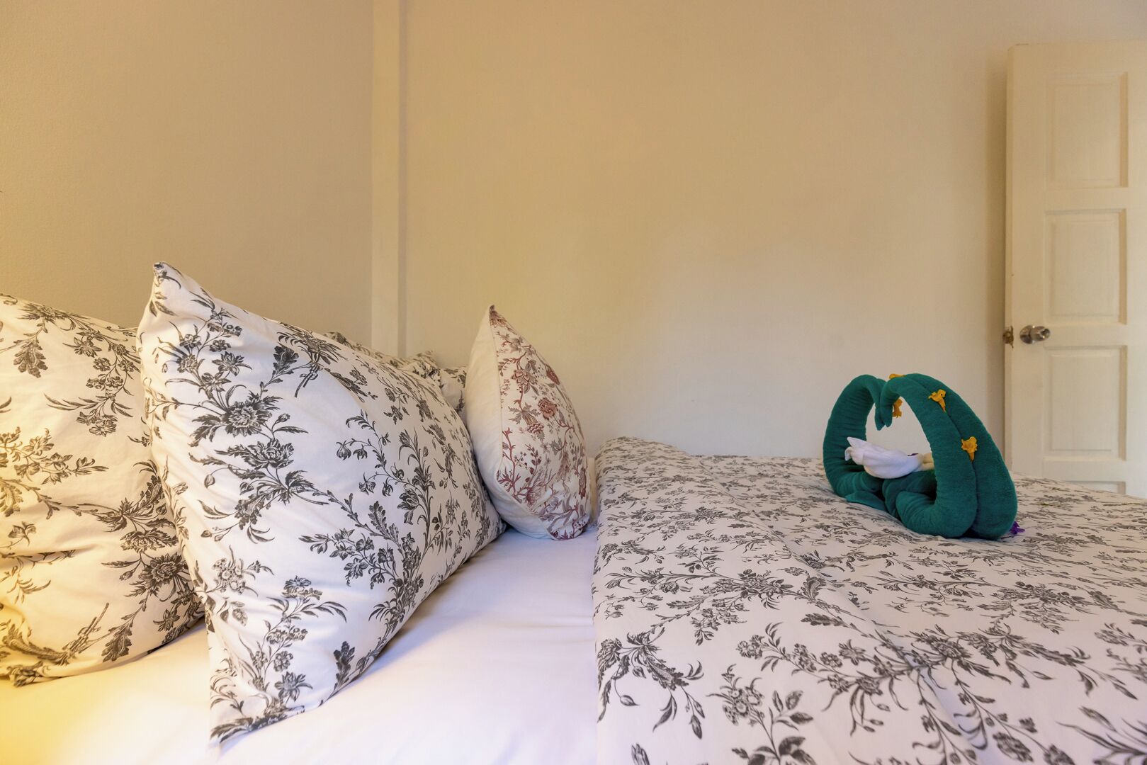 Ein Bett mit blumigem Bettzeug und Kissen, ein grünes Dekorationsobjekt in Form einer Schwan und eine weiße Tür im Hintergrund.