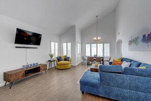 Een ruime woonkamer met een flatscreen televisie, een houten tv-meubel, een blauwe bank, een gele fauteuil, en een eethoek met een kroonluchter.