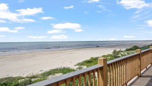 Vista a la playa desde una terraza de madera con barandilla, que da a una playa arenosa y el océano.