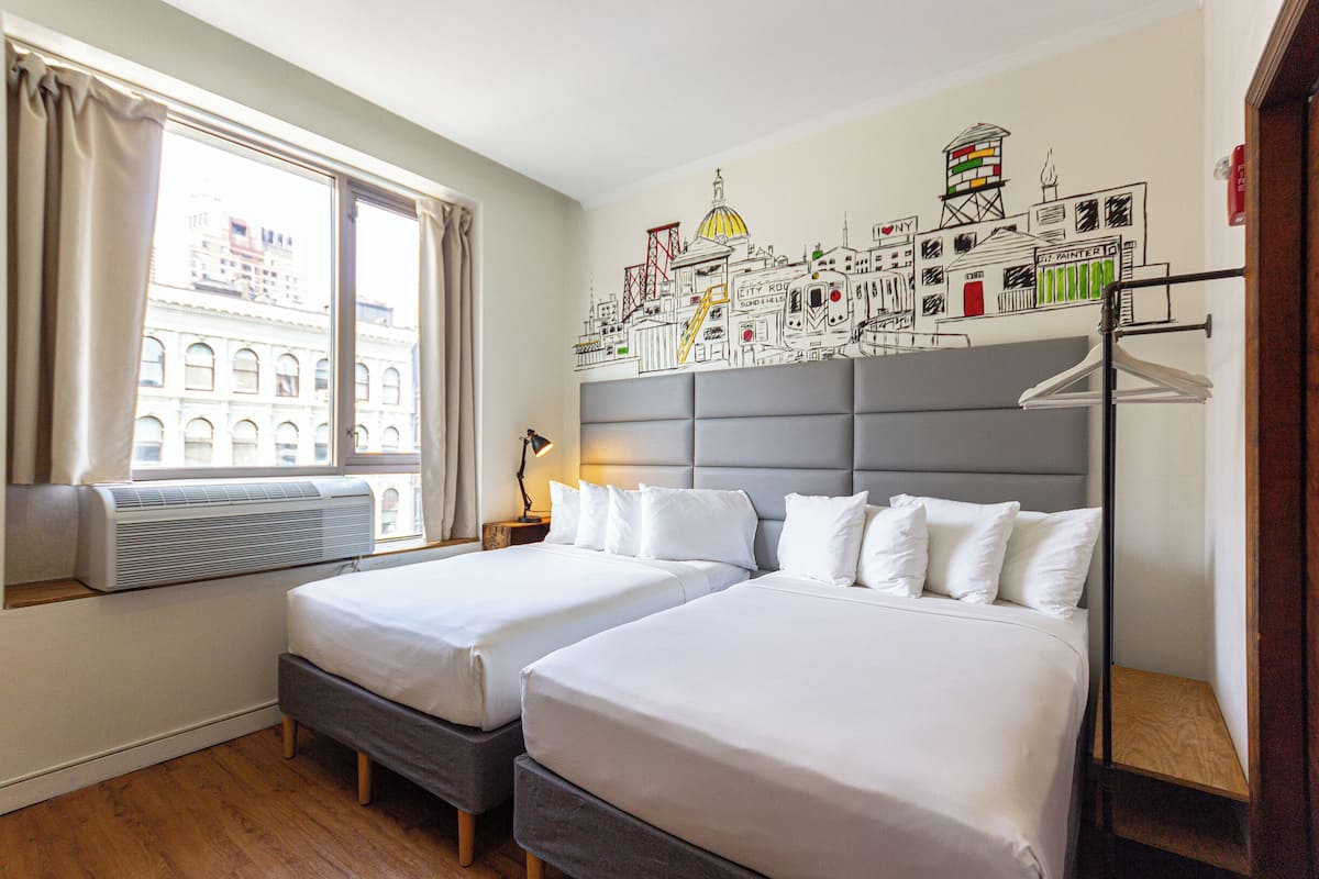 A hotel room with two beds, a large window with curtains, an air conditioning unit, and a cityscape mural on the wall.