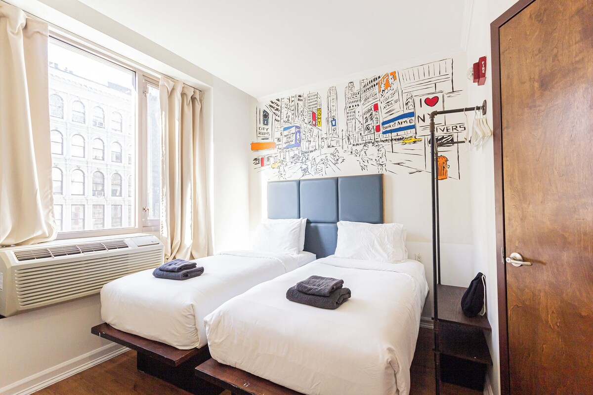 A hotel room with two beds, a large window with curtains, a wall mural, and a wooden door.