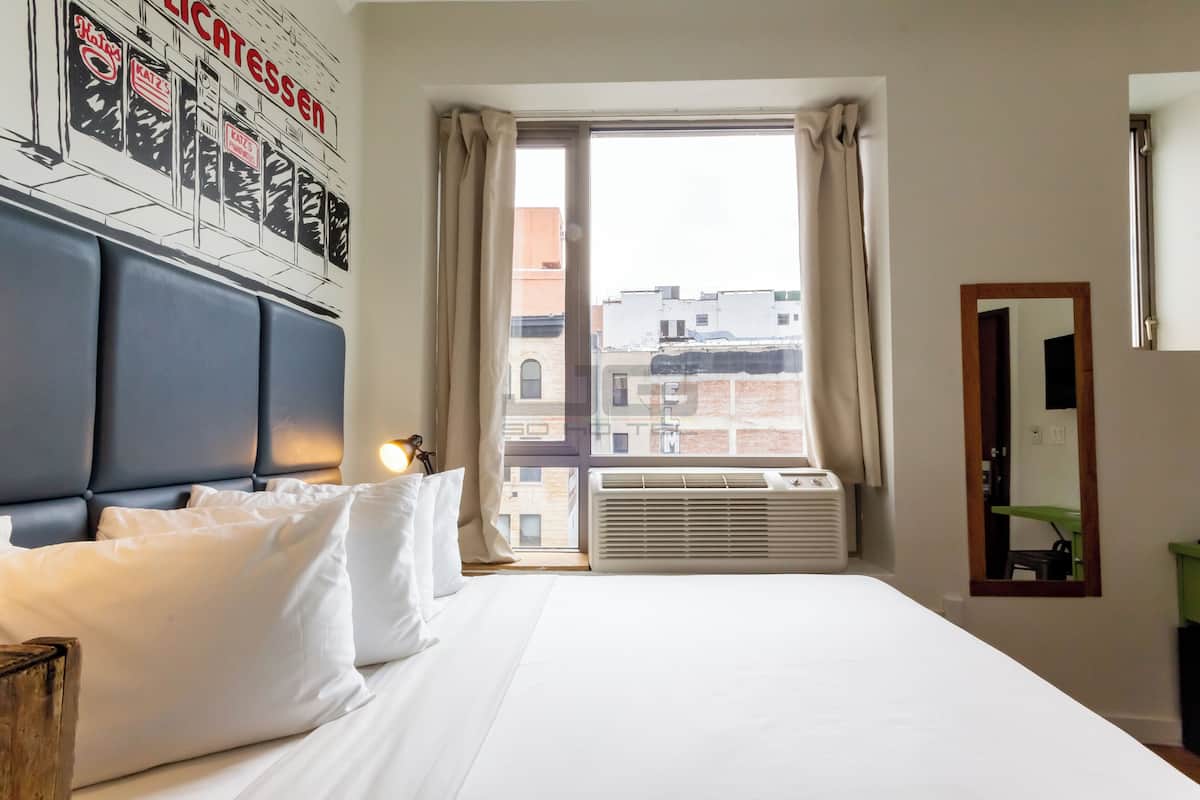 A hotel room with a large bed, a headboard, a window with curtains, an air conditioning unit, and a wall-mounted mirror.