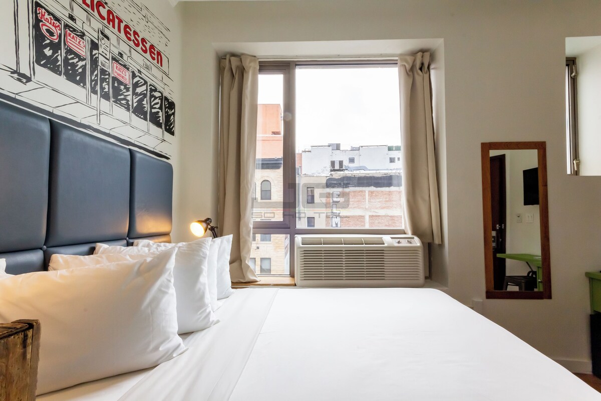 A hotel room with a large bed, a headboard, a window with curtains, an air conditioning unit, and a wall-mounted mirror.