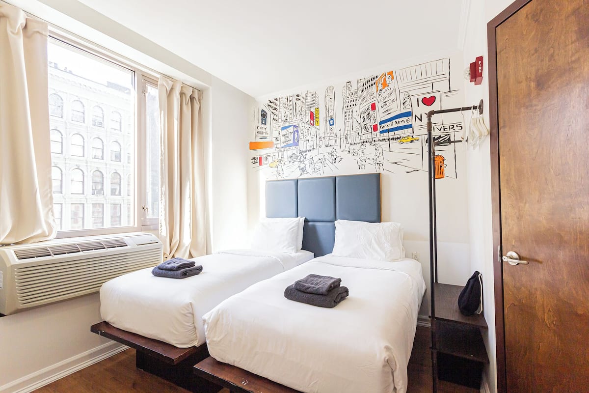 A hotel room with two beds, a large window with curtains, a wall mural, and a wooden door.