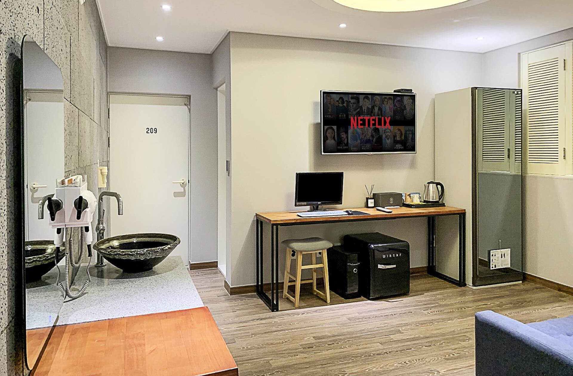 A modern hotel room with a wooden desk, a computer, a TV mounted on the wall, a large mirror, and a wardrobe with louvered doors.