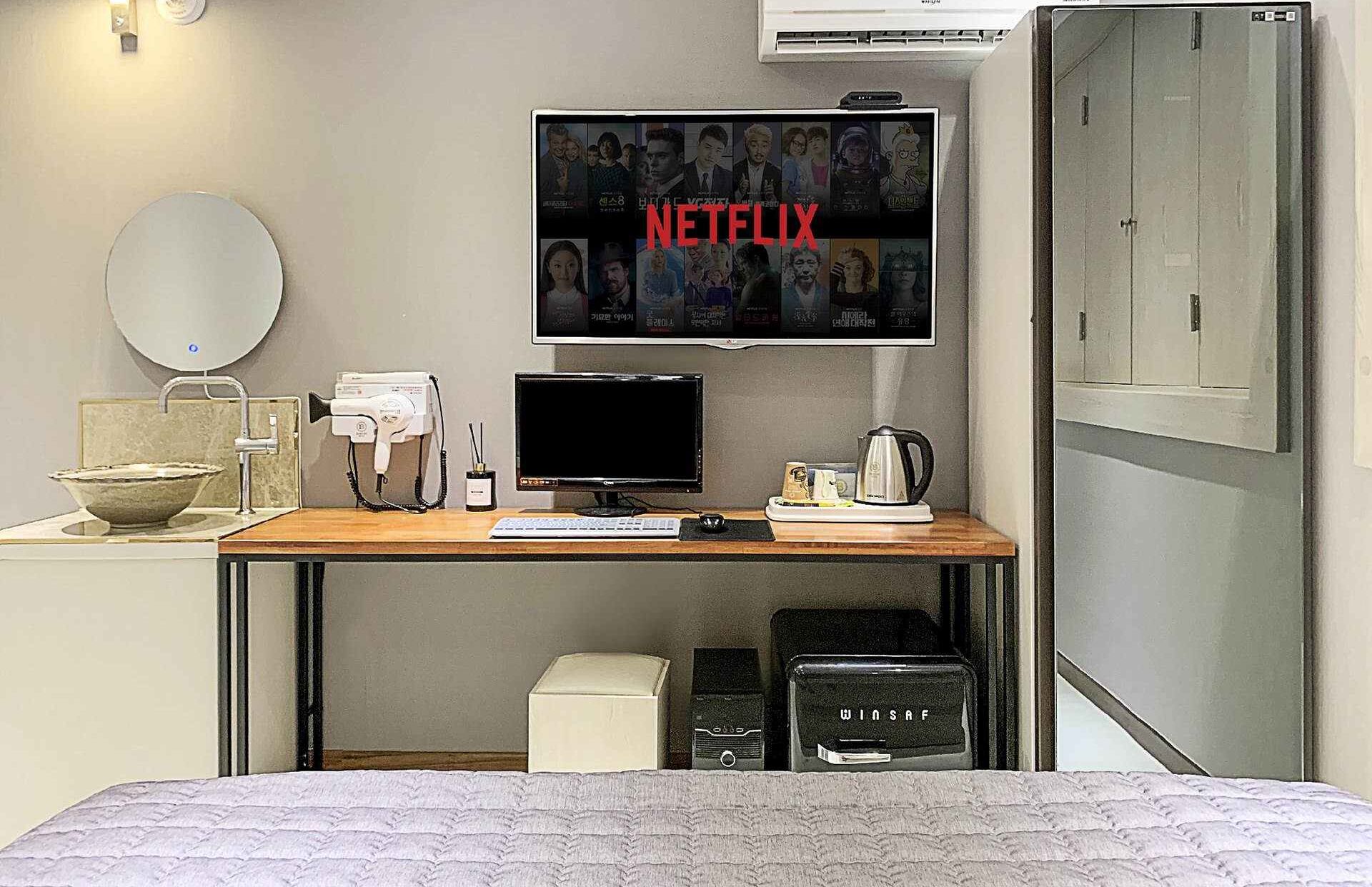 Hotellihuone, jossa on sänky, työpöytä tietokoneella, televisio, jossa näkyy Netflix, hiustenkuivaaja, vedenkeitin ja minibaari.