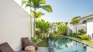 A modern outdoor pool area with a wooden deck, lounge chairs, and lush tropical plants. - Villa Casa Cielo by OriVista (Canggu)