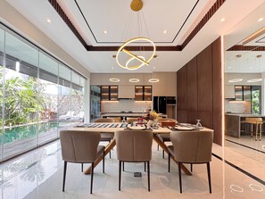 A modern dining area with a glass pool view, a central table with chairs, and a kitchen in the background. - Villa Louis Legian by Nagisa Bali (Legian)